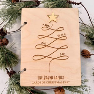 Puede incluir: Un tarjetero navideño de madera con un diseño de árbol. El árbol está hecho de líneas con los nombres Matías, Nicolás, Mamá y Papá. Una estrella dorada está en la parte superior. El texto "The Barni Family" y "Cards of Christmas Past" está en la parte inferior.