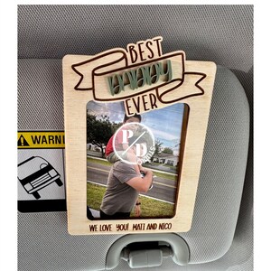 Puede incluir: Un marco de fotos de madera con el texto "Best Daddy Ever" y una foto de un padre y un hijo. El marco tiene una ranura para una visera de coche y el texto "We love you! Matt and Nico" está grabado debajo de la foto.