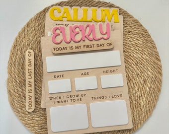 Puede incluir: Un letrero de madera con el texto "TODAY IS MY FIRST DAY OF" y espacios para la fecha, la edad, la altura, lo que el niño quiere ser cuando crezca y las cosas que le gustan. El letrero está decorado con los nombres "CALLUM" y "EVERLY" en letras amarillas y rosas.