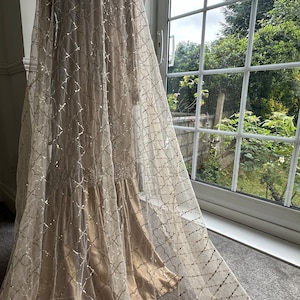 May include: A beige dress with a sheer overlay. The overlay features a geometric pattern of silver glitter. The dress is displayed near a window with a view of greenery.