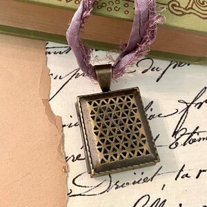 Puede incluir: Un colgante rectangular con acabado en bronce antiguo que presenta un patrón geométrico de recortes triangulares. El colgante está suspendido de una cinta de seda de color violeta claro deshilachada que sirve como cordón.