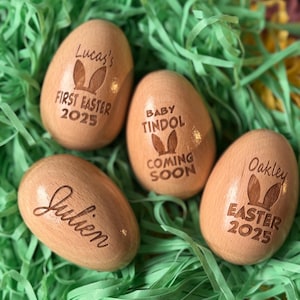 May include: Four wooden Easter eggs with bunny ears and personalized messages. The eggs are decorated with the names "Lucas", "Julien", "Oakley", and "Baby Tindol Coming Soon".