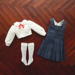 May include: A miniature school uniform set comprising a white blouse with a red bow, a navy blue pleated skirt dress, and white knee-high socks. The items are displayed on a dark wooden surface.