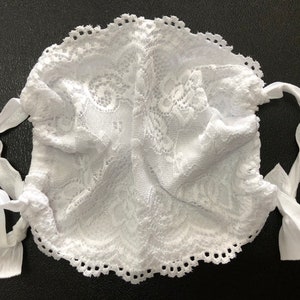 May include: White lace face mask with two white satin ties. The mask has a delicate floral pattern and a scalloped edge.