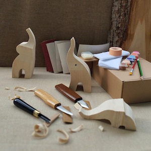 May include: A DIY wood carving kit featuring two standing light-colored wooden elephant figurines, one horizontal elephant shape, three carving knives with black, light wood, and dark wood handles, sandpaper sheets, paint pots, and brushes.