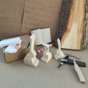 Könnte beinhalten: Ein Holzschnitzset mit drei unfertigen, hellen Holzfiguren in Tierform, einem braunen Pappkarton, weißen Stoffresten, einer Klebebandrolle, Buntstiften, einem kleinen Notizbuch, Farbtöpfen, einem Schnitzmesser und einer Holzscheibe.