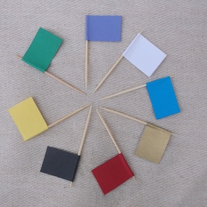 May include: Eight small flags of different colors are attached to toothpicks and arranged in a circle on a beige fabric background. The flags are yellow, green, blue, red, black, white, gold, and purple.