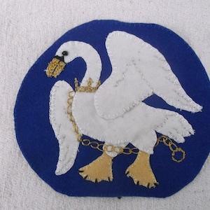 Puede incluir: Un cisne blanco con una cadena y una corona doradas, bordado en un círculo de fieltro azul.