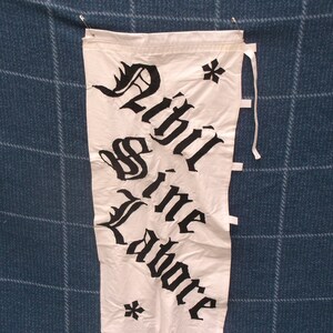 May include: A white fabric banner with black lettering that reads "Will the Labore" in a gothic font. The banner has two black stars at the top and bottom.