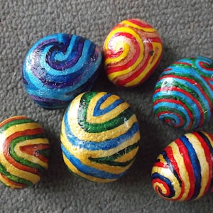 以下が含まれることがあります： 青、緑、赤、黄、金の色合いで渦巻き模様が描かれた、6つのペイントされた石。石は灰色の表面に円形に配置されています。