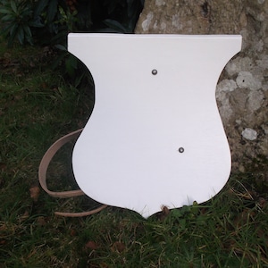 Puede incluir: Un objeto blanco de madera en forma de escudo con un diseño curvo, con dos pequeños remaches metálicos. Una correa de cuero marrón claro está sujeta al lateral. El objeto se encuentra sobre un fondo de hierba verde y un tronco de árbol.