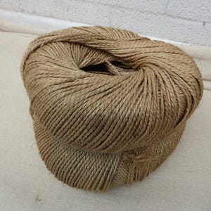 May include: A tightly wound ball of natural-colored twine. The twine is made of thick, twisted fibers, creating a textured surface. The ball is resting on a light-colored fabric, suggesting a craft or DIY project.