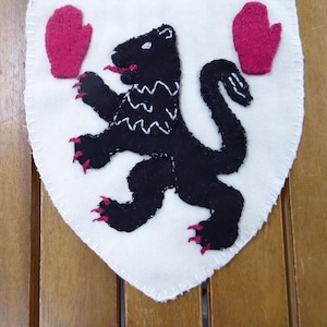 May include: A white felt shield featuring a black bear emblem with a red tongue and claws, and two red mittens. The bear has a white zig-zag pattern on its chest. The shield is stitched with white thread, set against a wooden background.