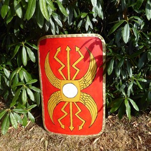 May include: A rectangular Roman-style scutum shield featuring a vibrant red background with a central gold-colored emblem. The design includes stylized golden wings and lightning bolt motifs surrounding a white circular center, finished with a gold trim.