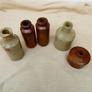 Peut inclure: Une collection de bouteilles antiques en grès et un couvercle. Les bouteilles varient en forme et en couleur, avec des nuances de marron, de beige et de beige. Une bouteille a un bec verseur, et une autre est arrondie. Le couvercle est de couleur marron foncé.