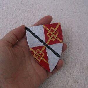 May include: A hand-painted shield with a heraldic design. The shield is divided into four sections: two silver and two red. Gold knotwork and a black diagonal line add detail. A unique, handcrafted item.