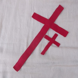 Puede incluir: Dos cruces rojas de diferentes tamaños se muestran sobre un fondo blanco. Las cruces parecen estar cortadas de tela o un material similar. La cruz más grande está colocada en diagonal, mientras que la más pequeña está horizontal.
