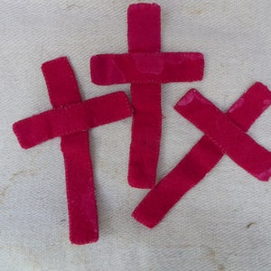 Puede incluir: Tres cruces de fieltro rojo, cada una con una forma y un tamaño diferentes. Las cruces están cosidas juntas y están sobre un fondo de tela blanca.