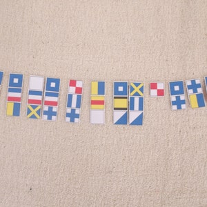 May include: A set of 14 nautical signal flags, each with a different colour and pattern. The flags are made of paper and are arranged in a row on a beige surface.