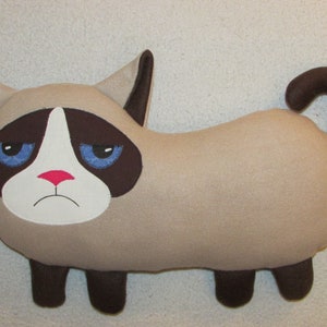 Könnte beinhalten: Ein Plüschtier eines mürrischen Katers mit braunem Fell, blauen Augen und einer rosa Nase. Die Katze liegt mit ihrem Schwanz hinter sich eingerollt.