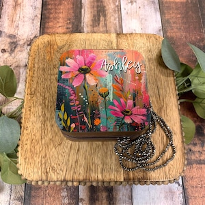 May include: A square, floral-patterned jewelry box with a zipper closure. The box has a colorful abstract design with pink flowers and the name "Ashley" written in white script. A silver necklace is lying next to the box.