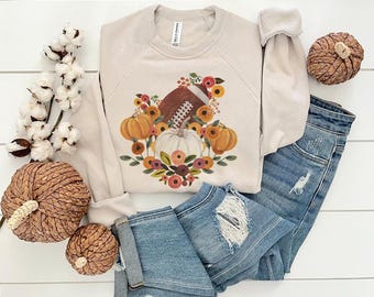 May include: A beige sweatshirt with a fall-themed graphic featuring a football, pumpkins, and floral accents. The image also includes a pair of denim shorts and decorative pumpkins, creating a seasonal aesthetic.