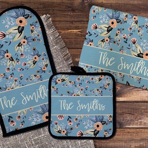 Op de afbeelding: Een bijpassende keukenset met een blauw bloemenpatroon en perzik- en crèmekleurige accenten. De set bevat een ovenwant, een pannenlap en een rechthoekige onderzetter. Elk item heeft de tekst "The Smiths" in een wit script.