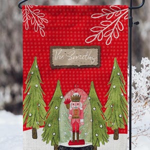 Puede incluir: Una bandera de jardín navideña roja con un patrón de lunares blancos y ramas blancas. La bandera presenta un cascanueces en una bola de nieve con dos pinos verdes a cada lado. El texto "The Smiths" está en una etiqueta de arpillera marrón.