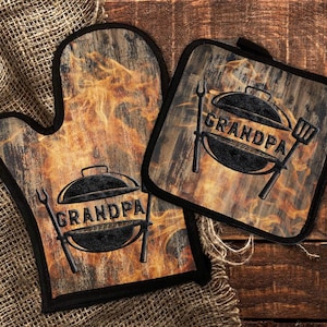 May include: A matching oven mitt and pot holder set with a wood-grain design and the word "GRANDPA" above a grill graphic. The background has a flame-like pattern. The set is trimmed in black.