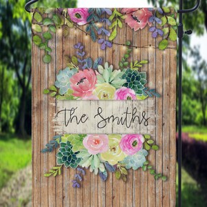 May include: A garden flag with a wooden plank background, adorned with watercolor flowers in pink, yellow, and green. The flag features the text "The Smiths" in elegant script, with string lights and greenery accents.