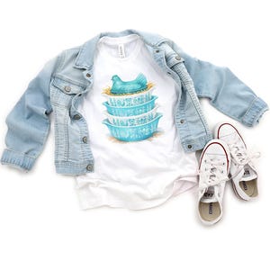 May include: A white t-shirt featuring a stack of turquoise bowls with a hen graphic. A light blue denim jacket and white sneakers with red accents are also in the image. The shirt has a classic, casual style.