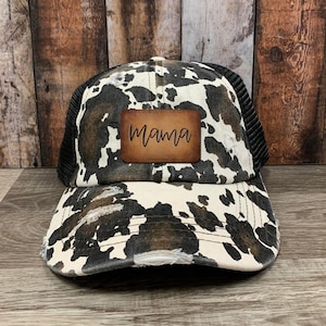 May include: A distressed trucker hat with a cow print design in black, white, and brown. The hat features a black mesh back and a brown leather patch with the word "Mama" in a script font.
