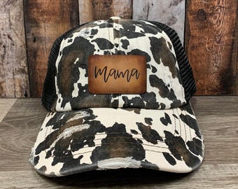 Könnte beinhalten: Distressed Trucker-Cap mit Kuhfellmuster in Schwarz, Weiß und Braun. Die Kappe hat eine schwarze Mesh-Rückseite und einen braunen Lederaufnäher mit dem Wort "Mama" in Schreibschrift.