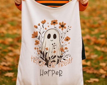 May include: White Halloween trick-or-treat bag featuring a friendly ghost design surrounded by orange flowers. The words "Trick or Treat" and the name "Harper" are printed on the bag. The bag is held by a person.