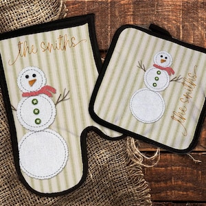 Puede incluir: Un juego de dos agarraderas de olla a rayas verdes y blancas con un diseño de muñeco de nieve. La agarradera de olla más grande tiene el texto "the Smiths" y la agarradera de olla más pequeña tiene el texto "the Smiths".