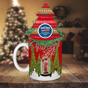 Könnte beinhalten: Eine weiße Keramiktasse mit einem Weihnachtsbaumdesign mit einem Nussknacker und dem Text "Ashley". Die Tasse ist mit einer roten und grünen Weihnachtsbaum-förmigen Schachtel mit einem Swiss Miss Heißgetränkepulver-Paket verziert.