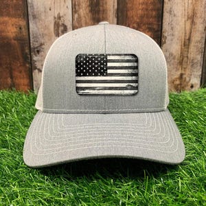 May include: A gray and white trucker hat with a distressed black and white American flag patch on the front.