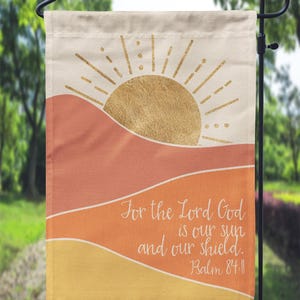 May include: A decorative garden flag with a warm color palette. The design features a golden sunburst over orange and yellow layered shapes. The text "For the Lord God is our sun and our shield. Psalm 84:11" is written in white.