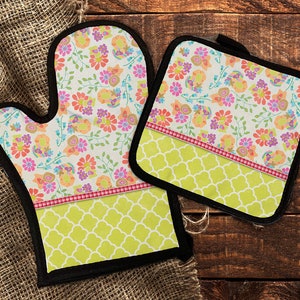 May include: A set of two oven mitts with a floral pattern. The mitts are green, pink, orange, and white. The mitts have a black trim and a green and white checkered pattern on the bottom.