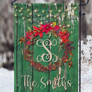 May include: A green wooden garden flag with a red poinsettia wreath and a gold letter "S" in the center. The wreath is decorated with red berries and green leaves. The flag also has a string of white lights and the text "The Smiths" at the bottom.