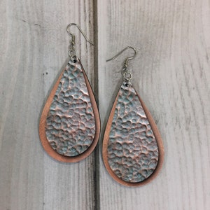 May include: A pair of teardrop-shaped earrings with a hammered copper finish. The earrings are made of two layers of leather, with the top layer being a lighter shade of copper than the bottom layer.