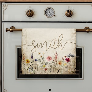 May include: A white kitchen towel with a floral design and the word "Smith" written in gold script. The towel is hanging on a metal rack above a white oven.