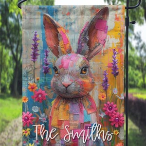 Puede incluir: Una bandera de jardín colorida con un conejo de retazos con un diseño abstracto y caprichoso. La bandera tiene un fondo brillante con flores y el texto "The Smiths" en una fuente blanca de escritura.