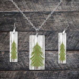 May include: A set of Christmas tree jewelry featuring a necklace and earrings. The pendant and earrings are rectangular and have a whitewashed wood background with a green Christmas tree painted on them. The tree has red ornaments.