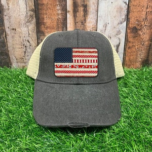 May include: A black and beige trucker hat with a distressed American flag patch. The flag is red, white, and blue with a paisley pattern.