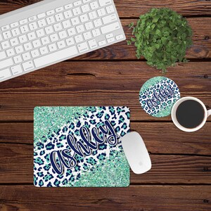 May include: A teal and navy blue mousepad with a glitter background and a leopard print design. The name "Ashley" is written in a white cursive font on the mousepad.