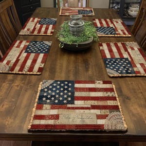 May include: A set of six rectangular placemats with an American flag design. The placemats feature a patchwork pattern with red and white stripes, a blue field of stars, and a decorative lace trim. The text "The Smiths" is visible on one placemat.