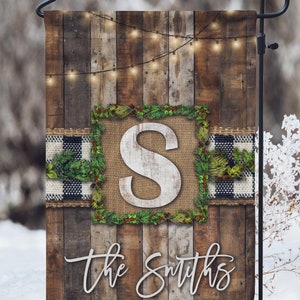 Puede incluir: Una bandera de jardín de Navidad de madera rústica con un fondo de arpillera y cuadros. La bandera presenta una gran letra blanca "S" rodeada de una corona de ramas de pino verdes y bayas rojas. El texto "The Smiths" está escrito en cursiva blanca en la parte inferior de la bandera.