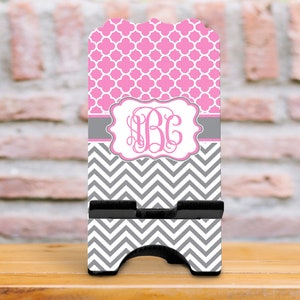 May include: A pink and gray chevron patterned phone stand with a monogrammed design. The monogram is in a white frame with a gray background. The phone stand is made of wood and has a black base.
