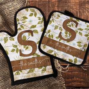 May include: Oven mitt and pot holder set with a floral design on a white wood background. The letter "S" and the name "the smiths" are in a brown wood tone. The set has a black border.